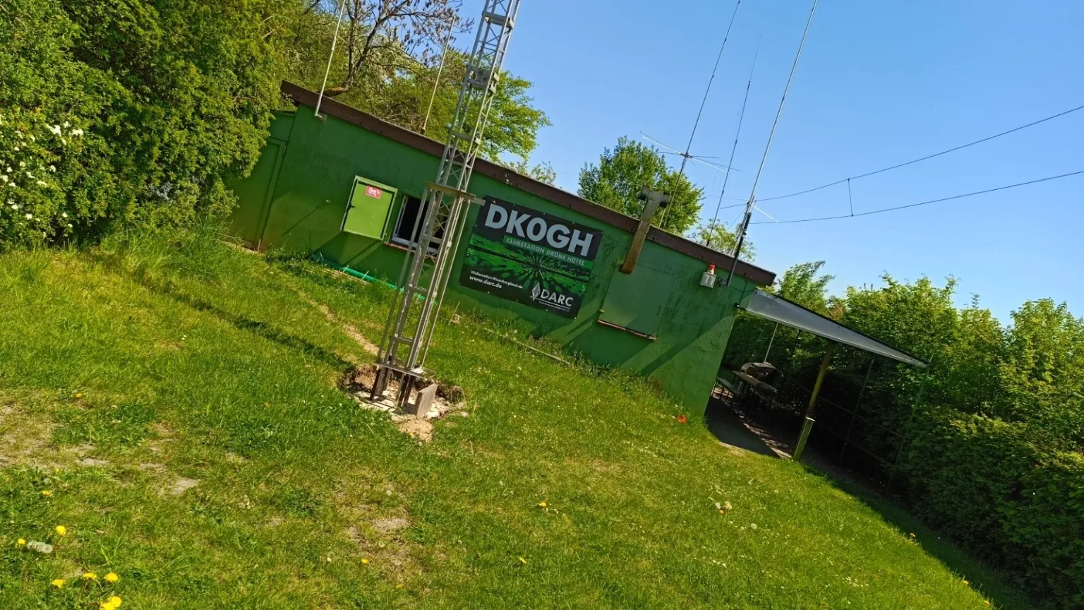 Seitliche Ansicht der grünen Clubstation DK0GH mit Antennen und einem Funkfreund am überdachten Sitzbereich